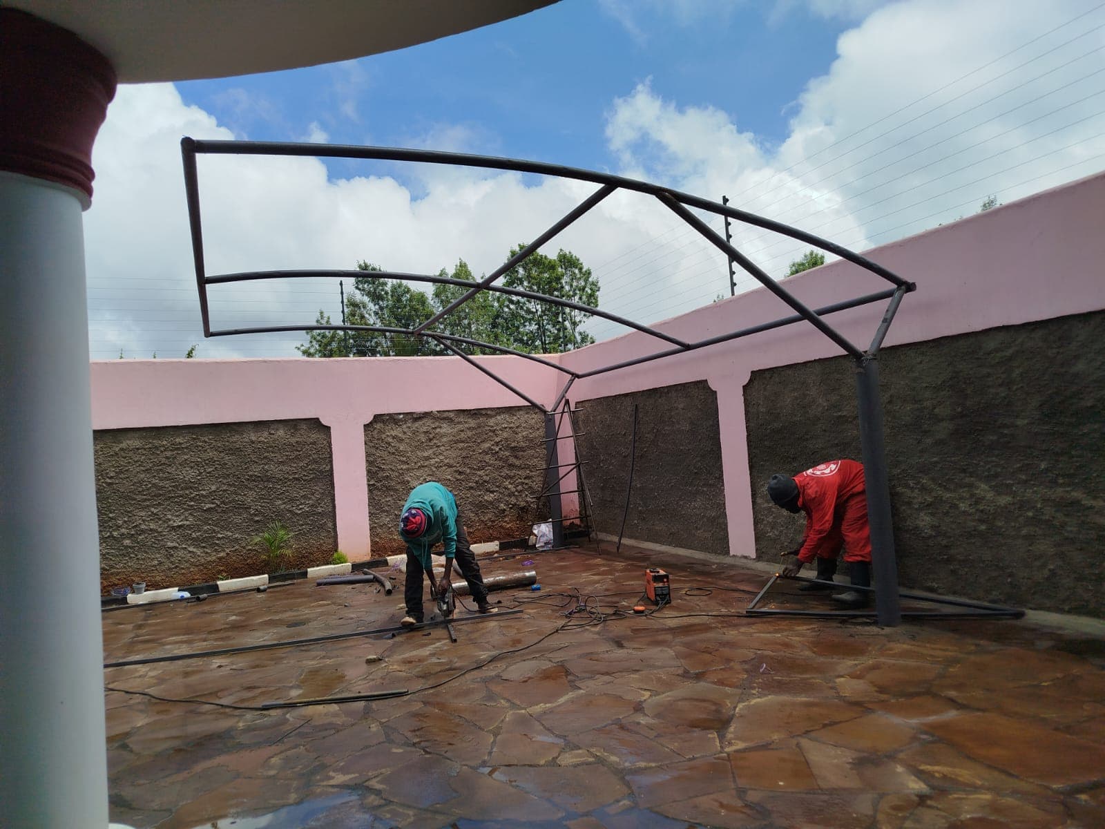 Two technicians welding a curved steel cantilever frame for a designer car parking shade at a Runda property