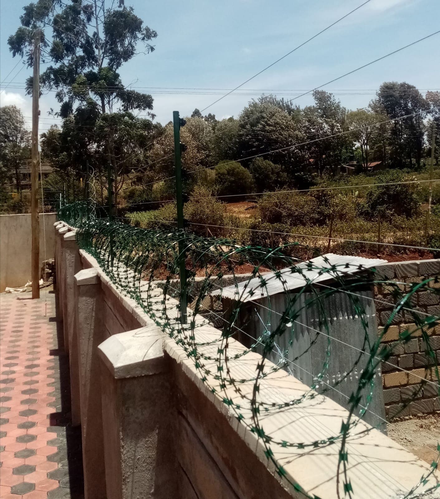 Green razor wire coils and electric fence wire strands installed along a stone-block boundary wall in Kikuyu, wide view