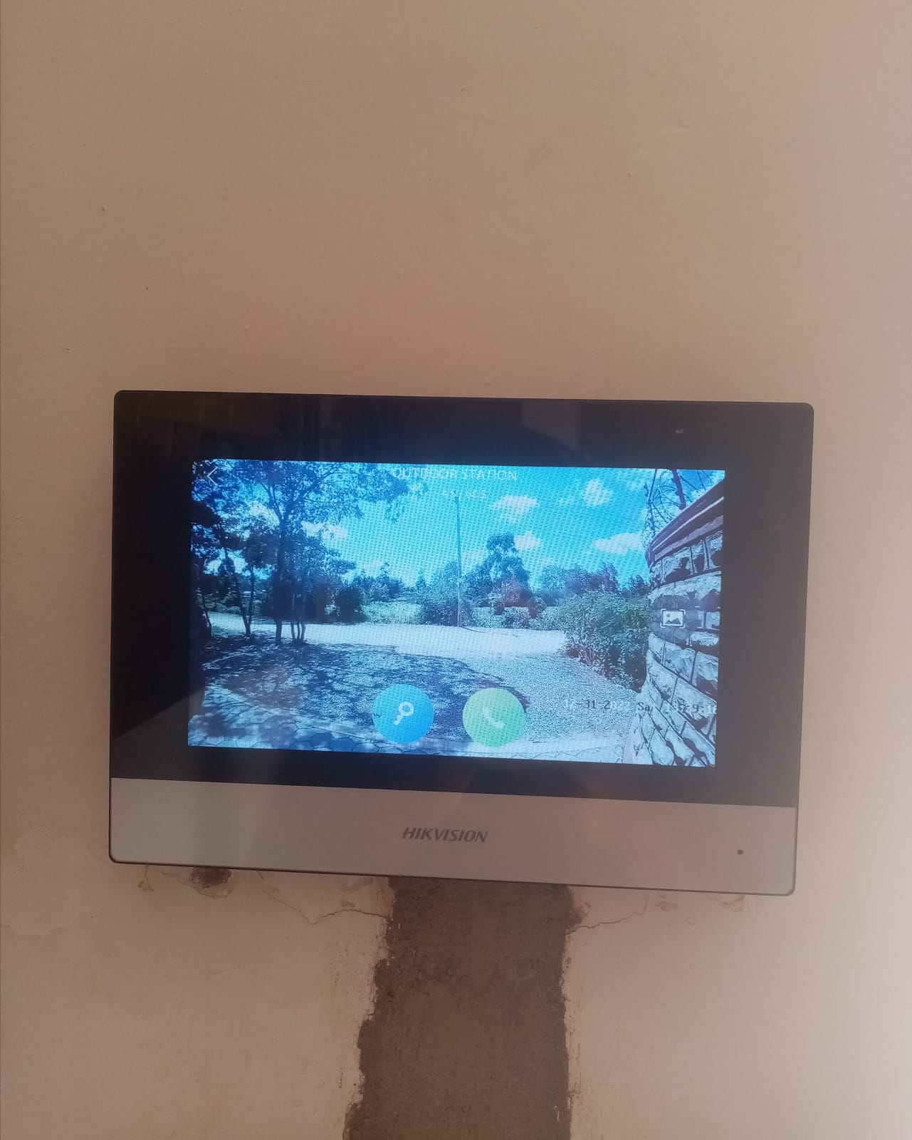 Hikvision indoor touchscreen video intercom monitor mounted on an interior wall, showing a live view of the outdoor station and driveway