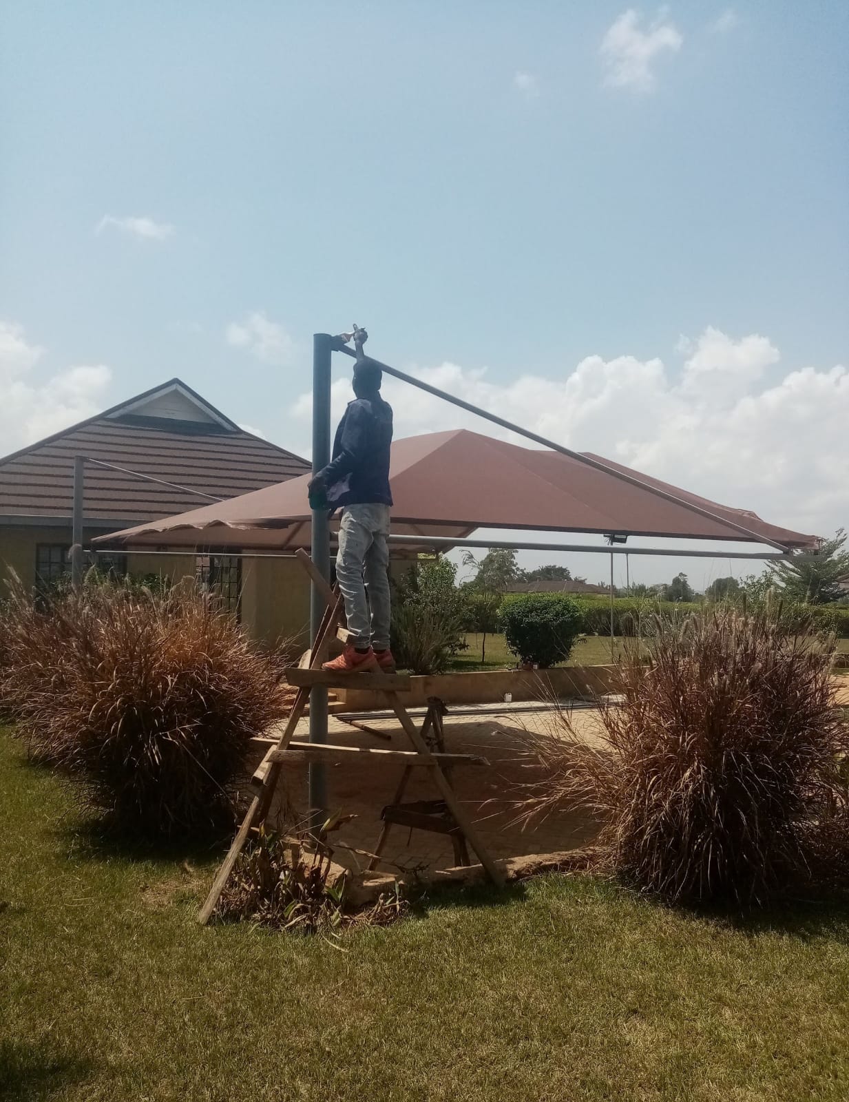 Technician on a ladder fitting the support post cap of a finished maroon car parking shade structure in Thika